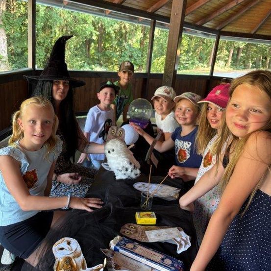 Eine Gruppe von Kindern und einer erwachsenen Frau sitzen um einen Tisch herum, wahrscheinlich in einem Wald. Die Frau trägt einen Hexenhut und hält eine Eule. Die Kinder lächeln und scheinen eine Aktivität zu machen. Der Tisch hat ein schwarzes Tuch, ein Glas, eine Schachtel und eine Papiereule. Der Hintergrund ist verschwommen mit Bäumen.