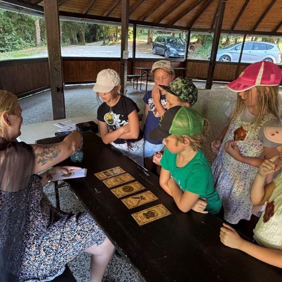 Eine Frau mit Tätowierungen liest Tarotkarten für eine Gruppe von Kindern in einem Pavillon. Die Kinder tragen Mützen und schauen aufmerksam zu.