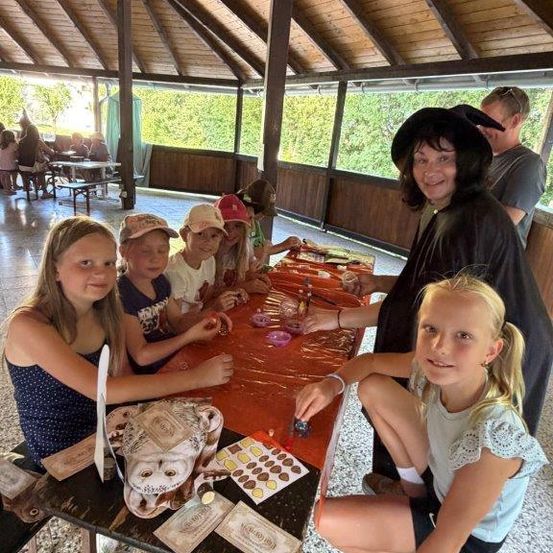 Eine Gruppe von Kindern und eine erwachsene Frau sitzen um einen Tisch in einem Pavillon herum und beschäftigen sich mit einer Bastelaktivität. Der Tisch ist mit einem orangefarbenen Tuch bedeckt und hat verschiedene Bastelmaterialien. Die Frau ist als Hexe gekleidet.