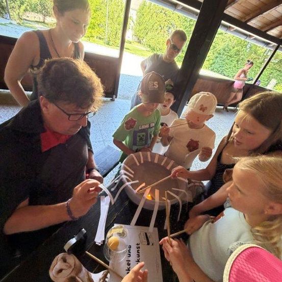 Eine Gruppe von Menschen, einschließlich Kindern, versammelt sich um einen Tisch in einem Park und röstet Marshmallows auf Stöcken über einem Feuer. Eine Frau mit Brille leitet die Aktivität.