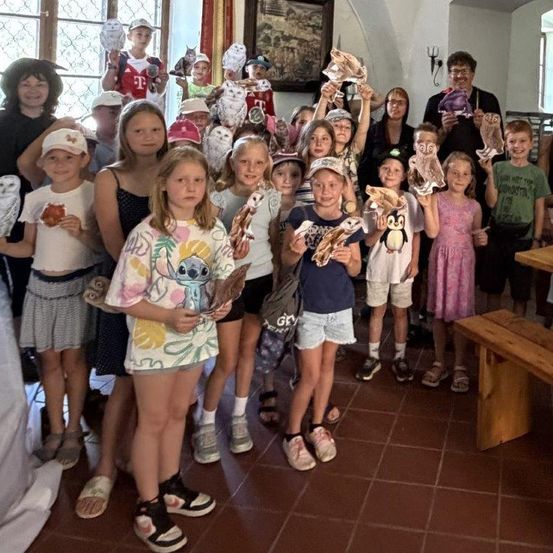 Eine Gruppe von Kindern und Erwachsenen posiert für ein Foto mit selbstgemachten Eulen-Basteleien in einem Raum mit einer Holzbank und gefliestem Boden.