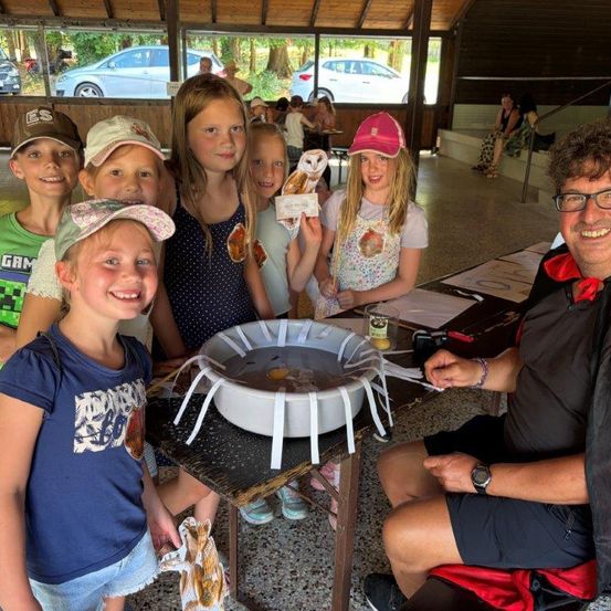 Eine Gruppe von Kindern und ein erwachsener Mann versammeln sich um einen Tisch mit einem Wasserbecken und einem Papierboot, lächeln und posieren für ein Foto.