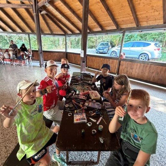 Eine Gruppe von Kindern sitzt in einem Pavillon um einen Tisch herum und ist in einer Bastelaktivität vertieft. Sie halten Pinsel in der Hand und haben verschiedene Bastelutensilien auf dem Tisch. Der Pavillon hat Holzwände und eine Decke. Im Hintergrund sitzen Menschen auf Bänken und ein Auto ist draußen geparkt.