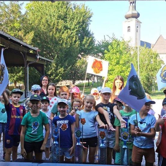 Eine Gruppe von Kindern in einem Park mit Flaggen und Auszeichnungen, mit einem Gebäude und Bäumen im Hintergrund.