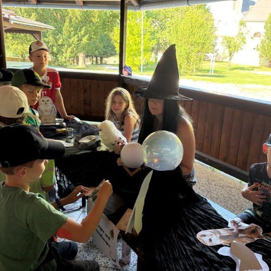 Eine Gruppe von Kindern und einer Frau in einem Hexenhut versammelt sich um einen Tisch mit einer weißen Eule und einem leuchtenden Ball. Sie befinden sich in einem Außenbereich mit einem Holzzaun und Bäumen im Hintergrund.