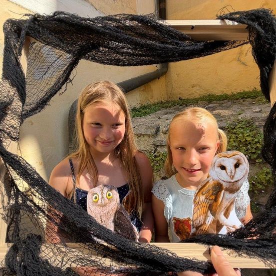 Zwei junge Mädchen lächeln und posieren für ein Foto mit Eulen-Ausschnitten in einem Rahmen. Der Rahmen besteht aus schwarzem Netz.