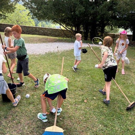 Eine Gruppe von Kindern spielt ein Spiel mit Besen und Bällen auf einer Grasfläche, mit einer Steinmauer im Hintergrund.