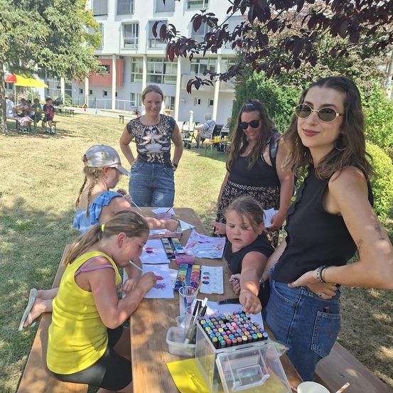 Eine Gruppe von Frauen und Kindern sitzt in einem Park um einen Tisch herum und beschäftigt sich mit einer Kunstaktivität. Der Tisch ist mit Kunstmaterial gefüllt, und die Frauen lächeln und schauen die Kinder an. Im Hintergrund sitzen Leute auf Bänken und ein Gebäude.