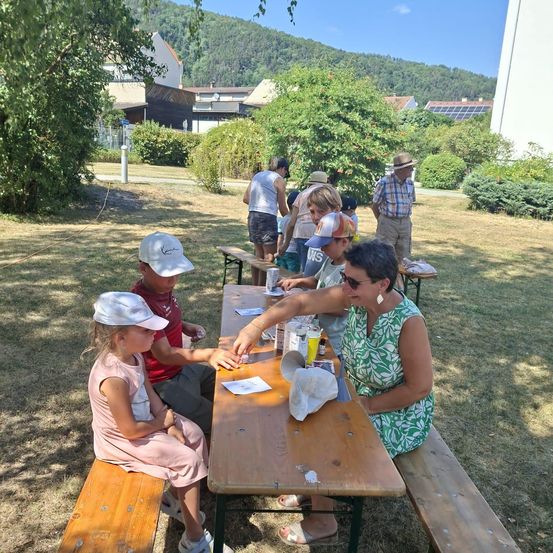 Eine Gruppe von Menschen, einschließlich Kindern, sitzt um einen Picknicktisch in einem Park und genießt Essen und Getränke. Im Hintergrund befinden sich Gebäude und Bäume.