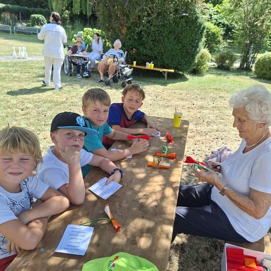 Eine Gruppe von Kindern und eine ältere Frau sitzen an einem Picknicktisch in einem Park, mit verschiedenen Bastelmaterialien auf dem Tisch. Im Hintergrund sitzen andere Menschen auf Bänken und eine Frau im Rollstuhl.