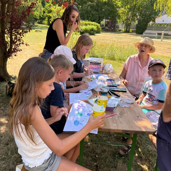 Eine Gruppe von Kindern und zwei Erwachsenen sitzt um einen Tisch in einem Park herum und ist in einer Malaktivität engagiert. Der Tisch ist mit Malbechern, Pinseln und Papier gefüllt. Die Kinder tragen Hüte und Sandalen.