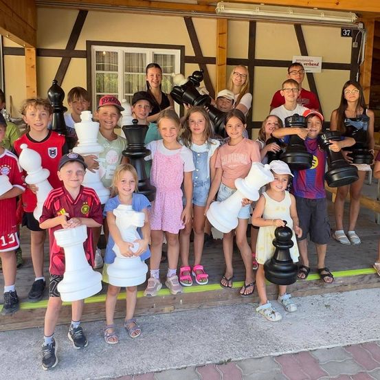 Eine Gruppe von Kindern und Erwachsenen posiert für ein Foto mit riesigen Schachfiguren vor einem Gebäude.