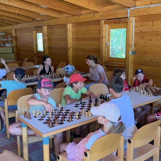 Eine Gruppe von Kindern und Erwachsenen spielt Schach in einem hölzernen Raum mit hölzernen Wänden und Tischen.