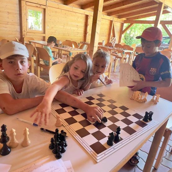 Vier Kinder spielen Schach an einem Tisch in einem hölzernen Raum mit hölzernen Wänden und Decke. Ein Kind hält ein Schachstück.