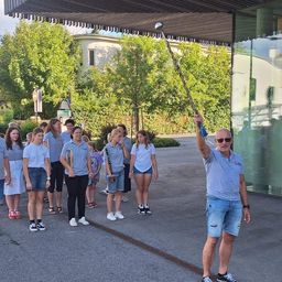 Eine Gruppe junger Menschen in blauen Shirts und Shorts, die Musikinstrumente halten, steht vor einem Gebäude mit einem Mann, der einen Taktstock hält.