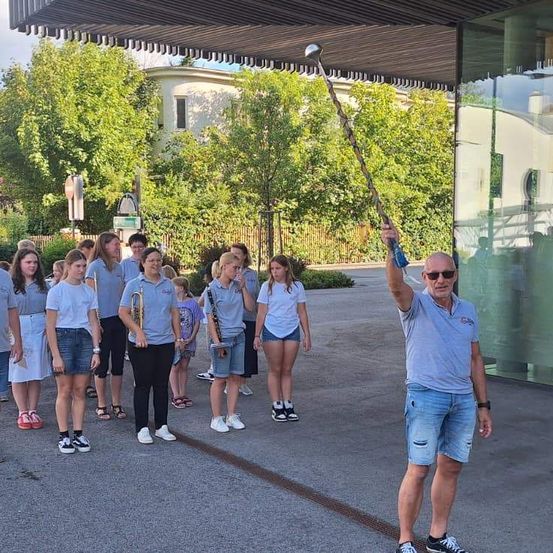 Eine Gruppe junger Menschen in blauen Shirts und Shorts, die Musikinstrumente halten, steht vor einem Gebäude mit einem Mann, der einen Taktstock hält.