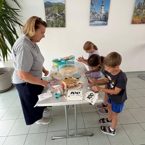 Eine Frau und drei Kinder verzieren Kekse an einem Tisch mit verschiedenen Gegenständen, darunter eine Schüssel mit Keksen und eine Schachtel mit der Aufschrift '100'.