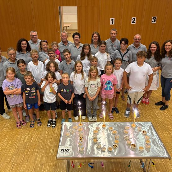 Eine Gruppe von Menschen, einschließlich Erwachsener und Kindern, versammelt sich um einen Tisch mit Cupcakes, um ein Ereignis zu feiern. Einige der Kinder halten Feuerwerkskörper. Auf dem Tisch steht die Nummer 100.