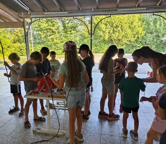 Eine Gruppe von Kindern und Erwachsenen versammelt sich um einen Tisch, möglicherweise für einen Workshop. Die Kinder halten Werkzeuge und scheinen an einer Aktivität beteiligt zu sein. Die Umgebung ist im Freien, mit Bäumen im Hintergrund.