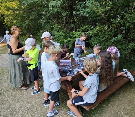 Eine Gruppe von Kindern und Erwachsenen versammelt sich um einen Picknicktisch in einem Park und beschäftigt sich mit einer Aktivität. Der Tisch ist mit verschiedenen Gegenständen bedeckt, und die Kinder tragen Mützen und Turnschuhe. Im Hintergrund befinden sich Bäume und ein Weg.