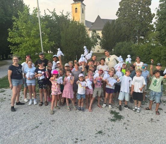 Eine Gruppe von Kindern und Erwachsenen posiert für ein Foto vor einer Kirche und hält weiße Objekte.