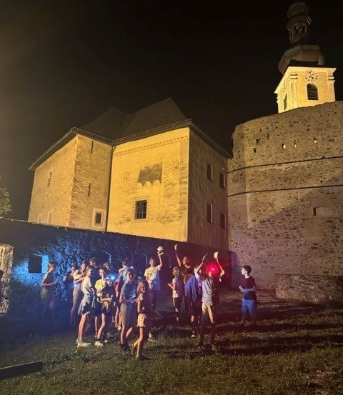 Eine Gruppe von Menschen versammelt sich bei Nacht vor einer historischen Burg, beleuchtet von Taschenlampen und blauen Lichtern.