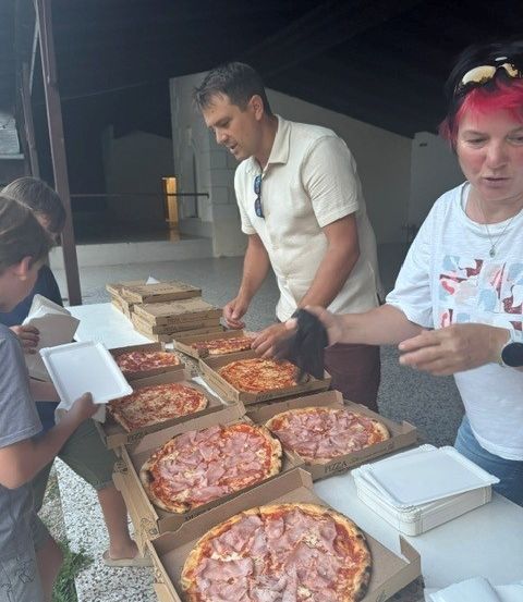 Eine Gruppe von Menschen steht um einen Tisch herum, der mit Pizzaschachteln gefüllt ist. Ein Mann hält eine Pizza, während eine Frau ihn ansieht.