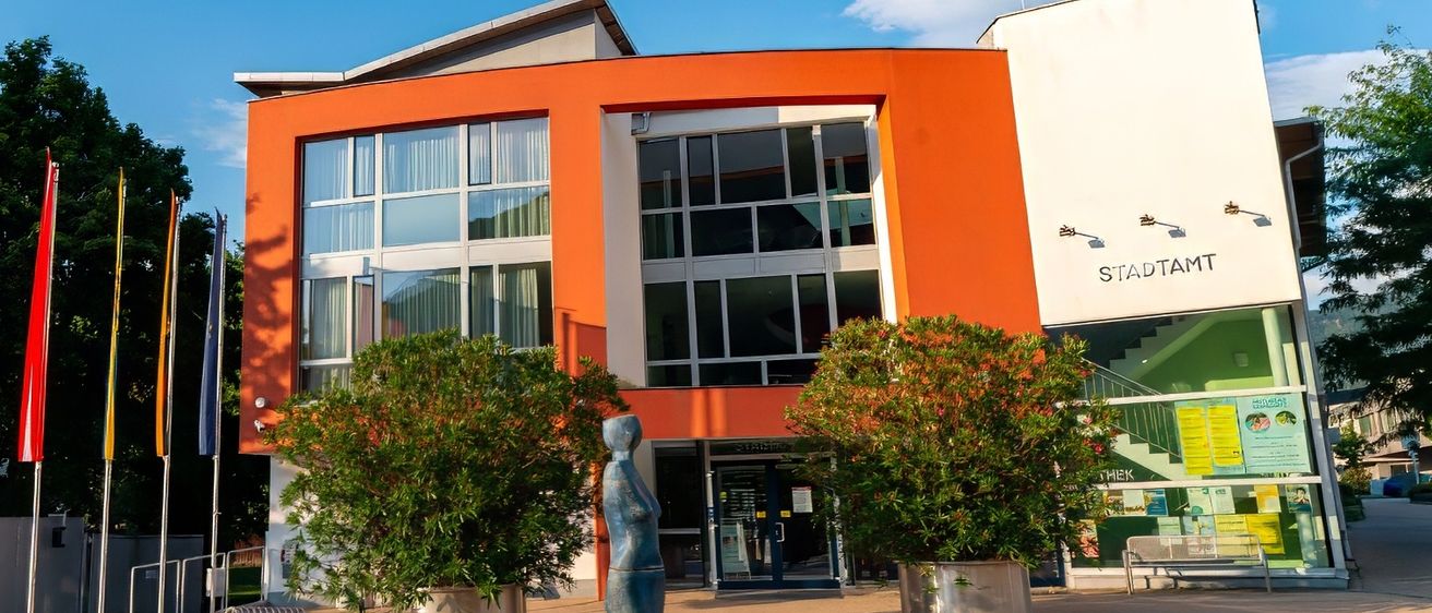 Ein modernes Gebäude mit orangefarbenen Wänden und großen Fenstern steht vor einem blauen Himmel. Zwei große Bäume und eine Statue sind davor. Das Gebäude hat das Wort Stadtamt darauf.