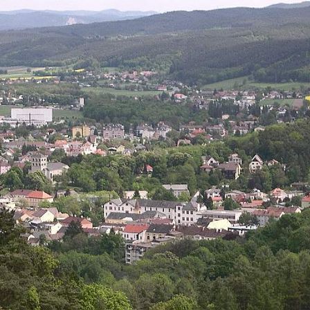 Ein Luftbild einer Stadt, umgeben von Bäumen und Bergen. Gebäude mit roten Dächern sind über das Gebiet verteilt.