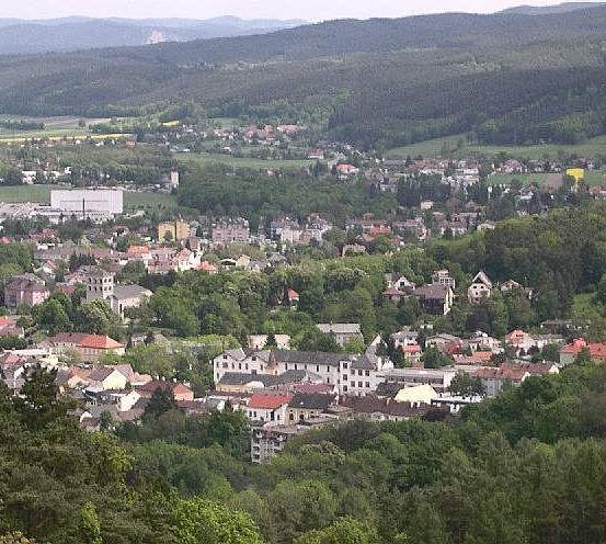 Ein Luftbild einer Stadt, umgeben von Bäumen und Bergen. Gebäude mit roten Dächern sind über das Gebiet verteilt.