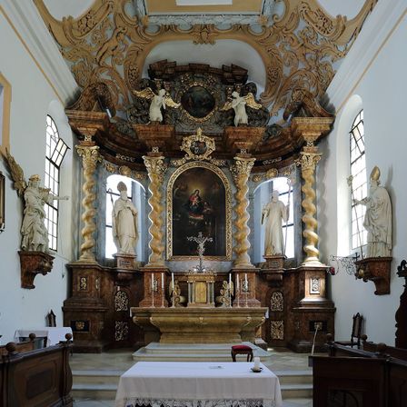 Der reich verzierte Altar einer Kirche zeigt ein großes Gemälde in einem goldenen Rahmen, flankiert von Statuen. Zwei Engel thronen auf dem Altar, und kunstvolle Holzarbeiten schmücken die Decke.