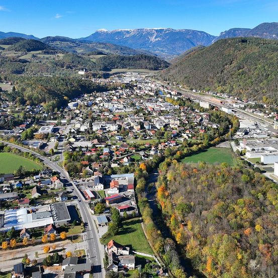 Luftaufnahme einer Stadt mit vielen Gebäuden, eingebettet zwischen Bergen und einem Fluss, mit grünen Bäumen und blauem Himmel.