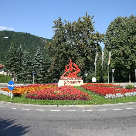 Ein Schild für die Einkaufsstadt Gloggnitz mit roten Blumen im Vordergrund und Bäumen im Hintergrund.