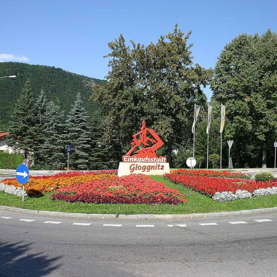 Ein Schild für die Einkaufsstadt Gloggnitz mit roten Blumen im Vordergrund und Bäumen im Hintergrund.