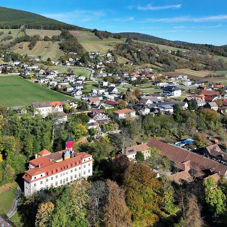 Luftaufnahme eines Dorfes mit verschiedenen Häusern, einem markanten weißen Gebäude mit rotem Dach und einer Kulisse aus Hügeln und Feldern unter einem klaren Himmel.