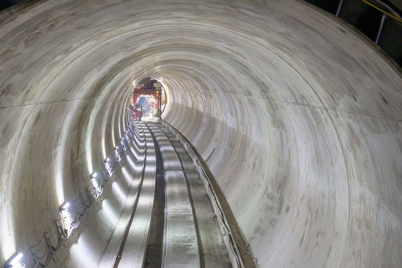 Ein langer, gewölbter Tunnel mit Betonwänden und Stahlschienen ist von innen zu sehen. Links sind Lichter und in der Ferne ein Baufahrzeug.