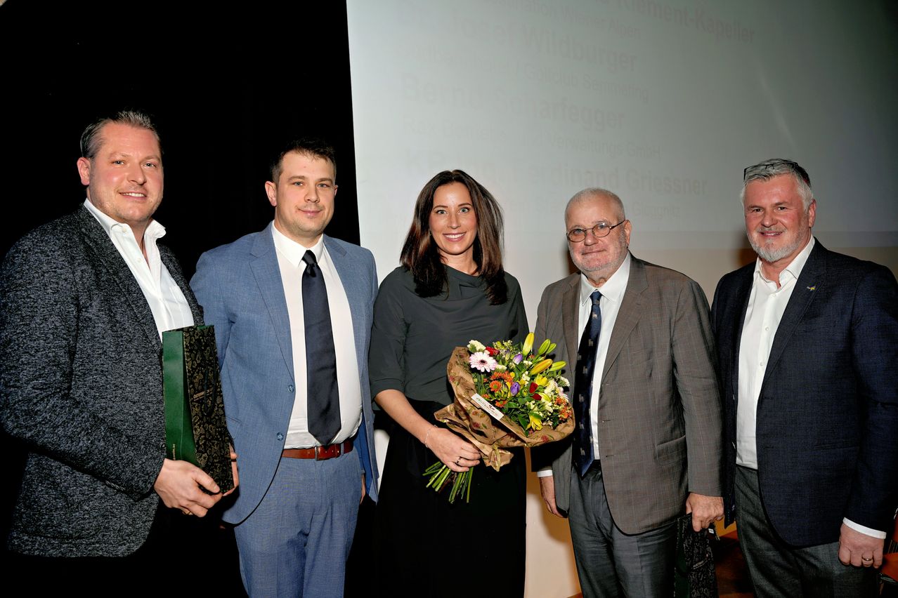 Fünf Personen stehen zusammen für ein Foto. Eine Frau in der Mitte hält einen Blumenstrauß. Hinter ihnen ist eine weiße Leinwand mit Text.