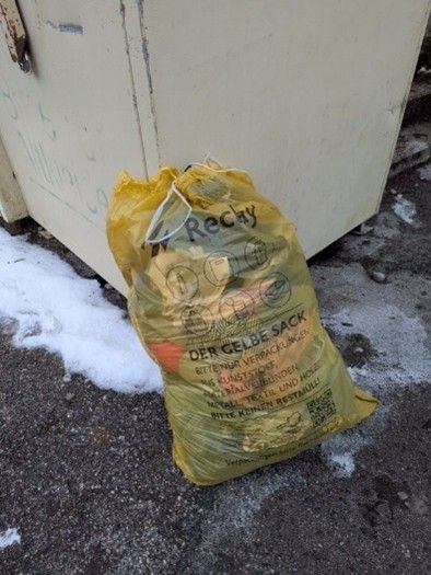 Ein gelber Sack mit der Aufschrift 'Der gelbe Sack' liegt neben einem weißen Container mit Graffiti auf dem Boden.