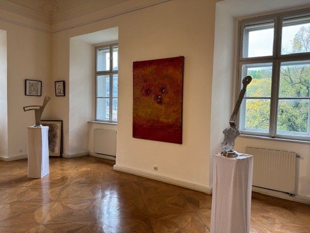 Ein leerer Raum mit zwei Skulpturen auf Podesten, einem großen Gemälde an der Wand, gerahmten Bildern und Fenstern mit Bäumen draußen.