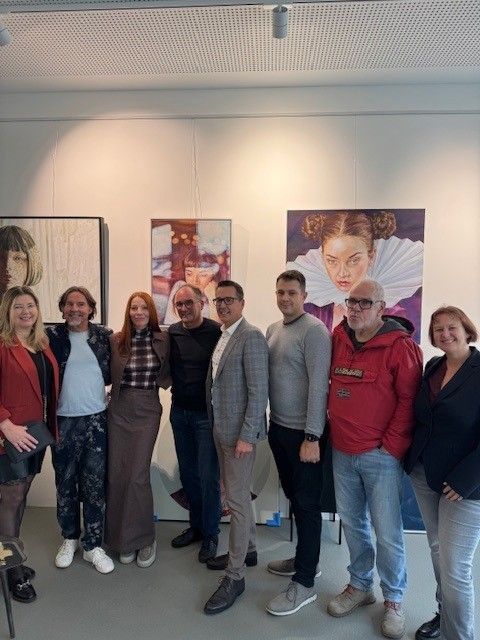 Eine Gruppe von Menschen steht in einer Kunstgalerie zusammen. Sie posieren für ein Foto mit verschiedenen Gemälden im Hintergrund.