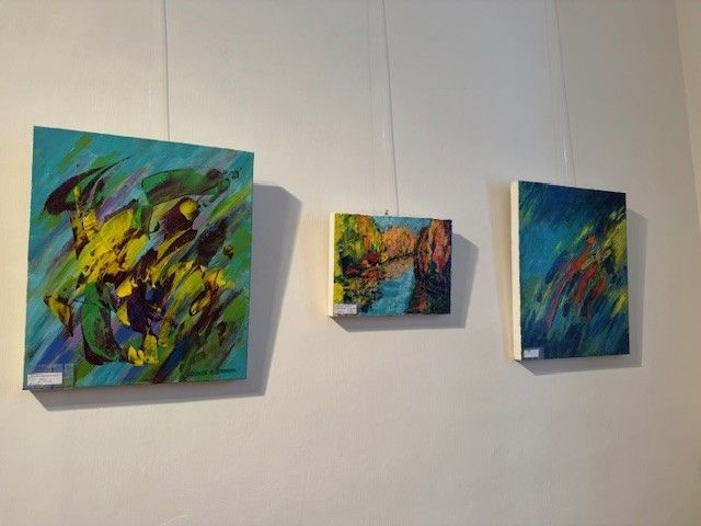 Drei abstrakte Gemälde hängen an einer weißen Wand. Das linke Gemälde ist groß und hat leuchtende gelbe und grüne Farben. Das mittlere Gemälde ist kleiner und zeigt eine herbstliche Landschaft. Das rechte Gemälde ist groß und zeigt eine Mischung aus blauen, roten und gelben Farben.