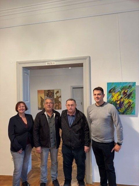 Vier Personen, zwei Männer und zwei Frauen, stehen gemeinsam in einer Kunstgalerie und lächeln für ein Foto. Sie stehen vor zwei Türen und Gemälden an den Wänden.