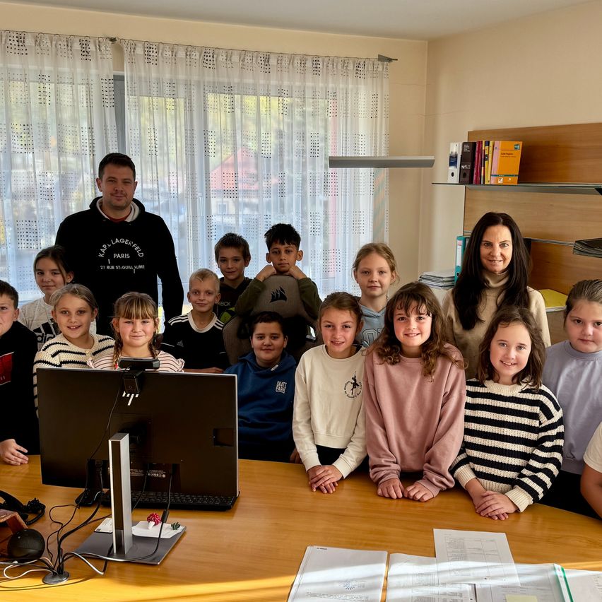 A group of children and two adults are posing for a photo in a classroom, with a monitor and desk in front of them. The classroom has a white curtain, shelves, and books.