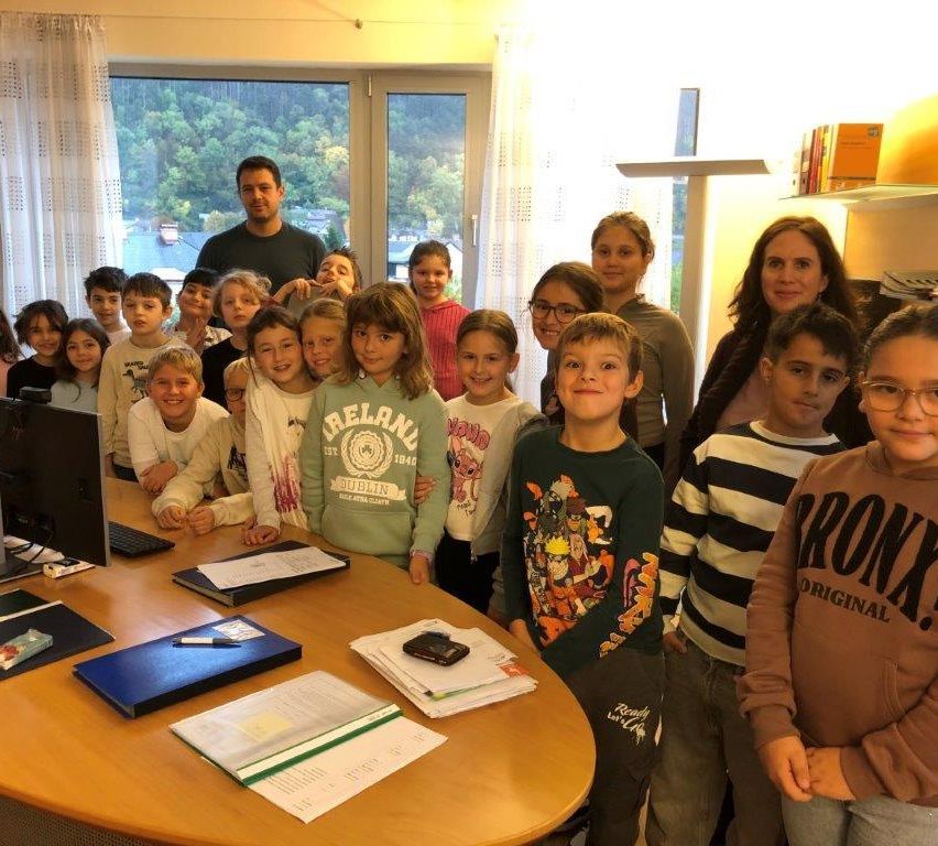 A group of children and adults stand in front of a window, smiling and posing for a photo. They are in a room with a table and books.