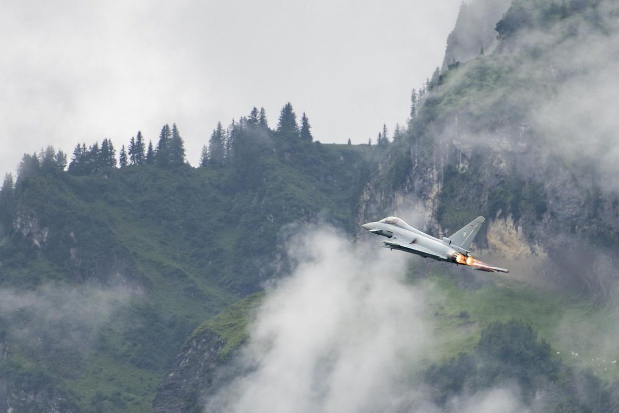 A fighter jet with afterburners on, flying between two mountains with trees on the top. The mountain range is foggy.