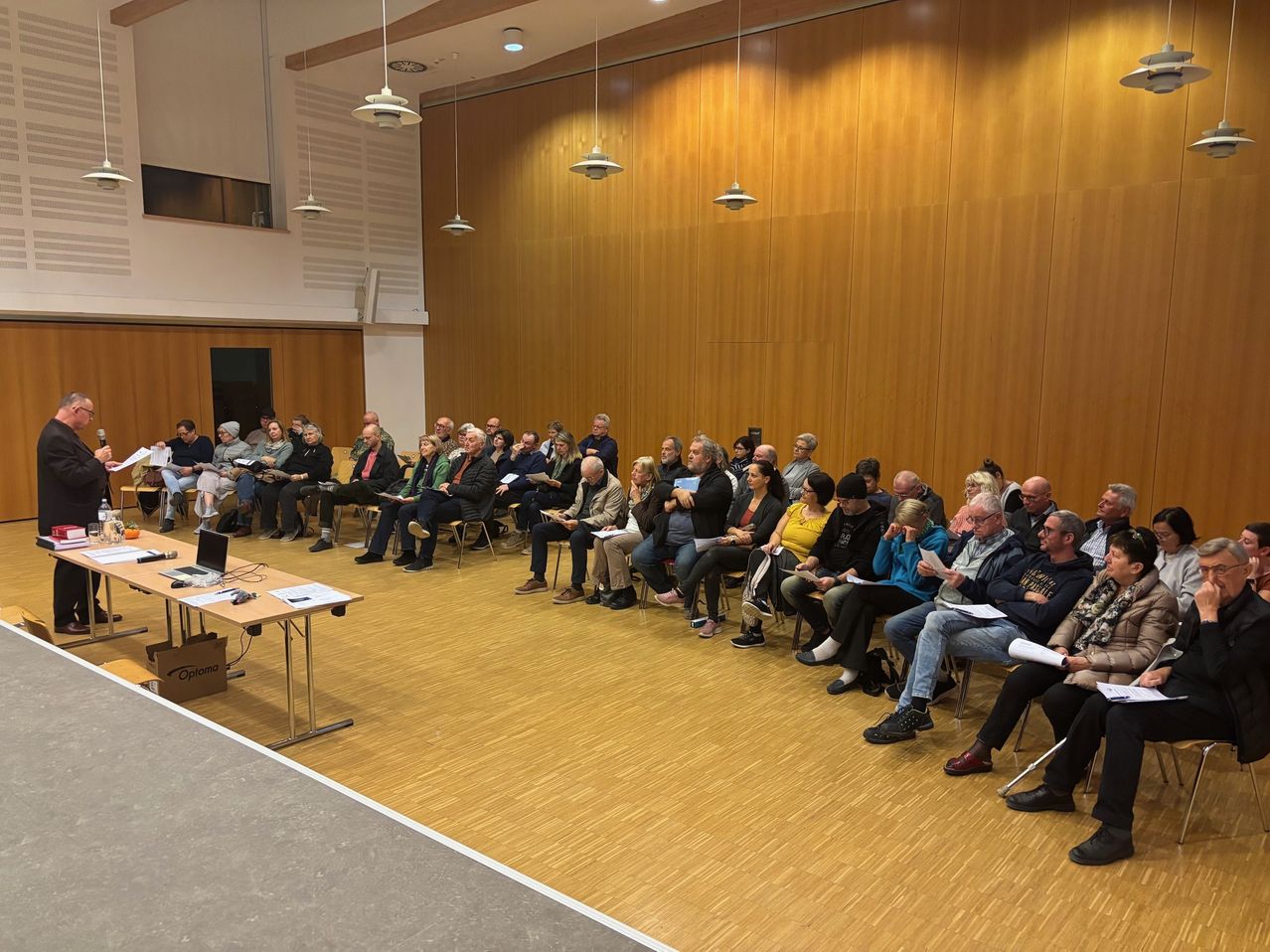 A group of people sit in chairs in a room with wooden walls. Some hold papers, and a laptop is on a table.