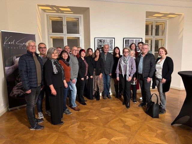 Eine Gruppe von Menschen steht zusammen in einem Raum, einige lächeln für ein Foto. Sie tragen Winterkleidung. Hinter ihnen wird ein Banner angezeigt. Der Raum hat Holzböden und weiße Wände mit gerahmten Bildern.