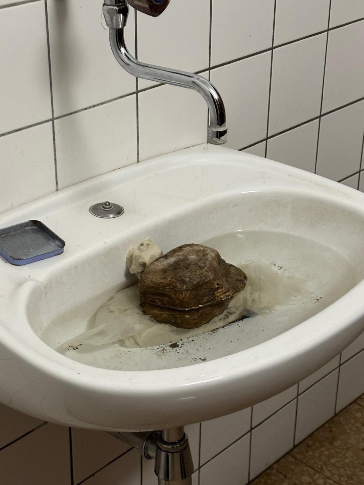 Ein weißes Badezimmerbecken mit einem Wasserhahn hält einen großen Felsen, in Stoff gewickelt, in Wasser eingetaucht, neben einem kleinen, blauen Tablett.
