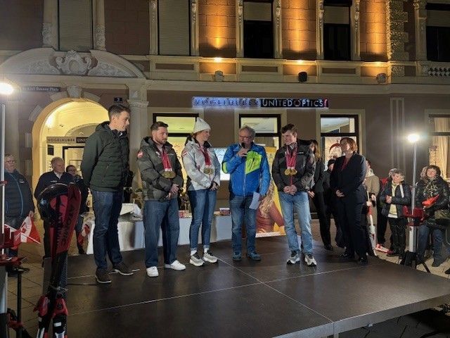 Five people stand on stage with medals. They are wearing winter clothes and smiling. A man holds a microphone, possibly giving a speech. A banner with a logo is behind them. The stage is on a city street.