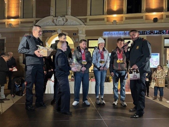 A group of people are standing on a stage, likely for an award ceremony. They are wearing jackets and sneakers, and some of them are wearing medals. One man is holding a trophy and a microphone.